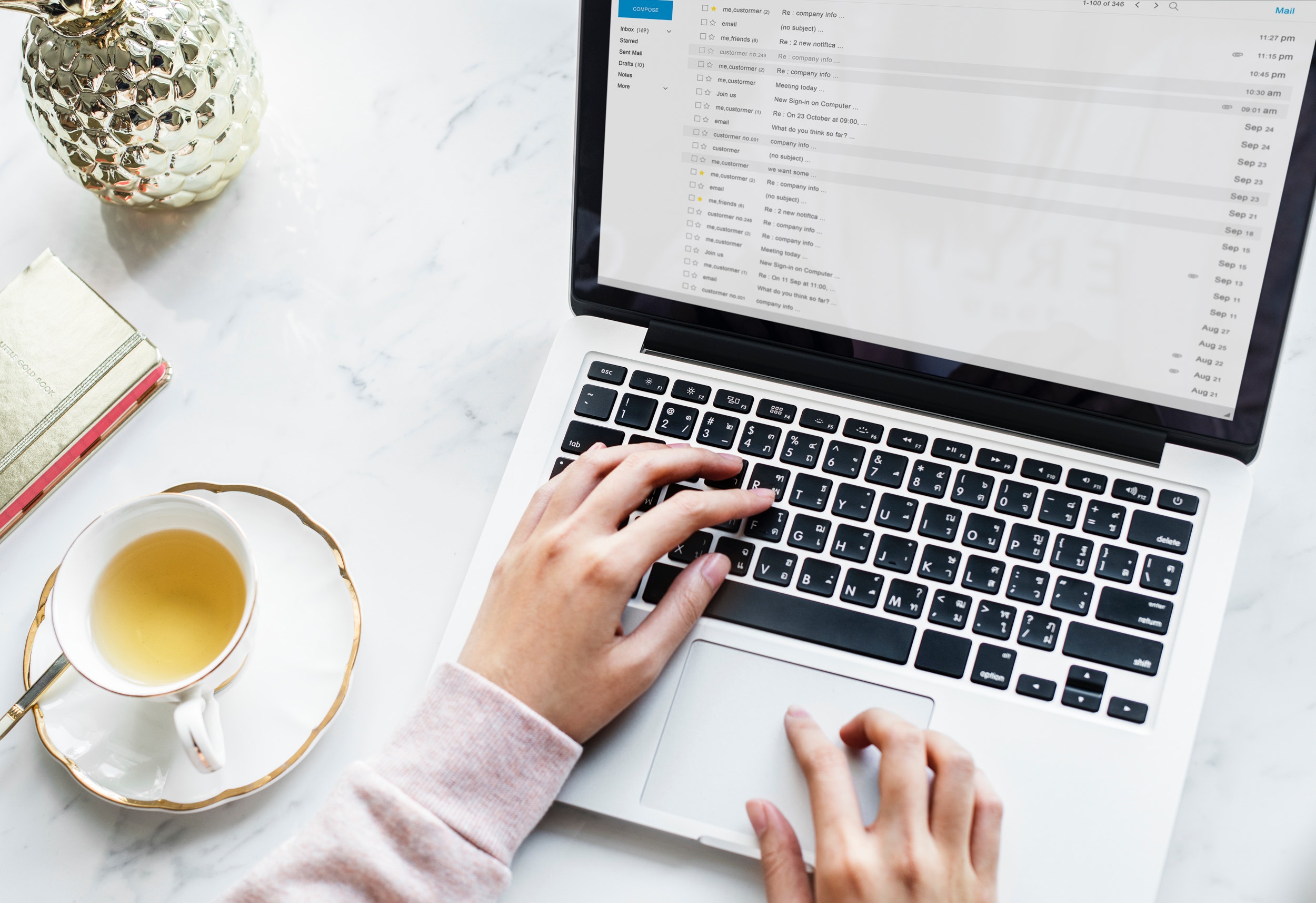 Mano escribiendo en laptop sobre mesa con taza de té y cuaderno decorativo.